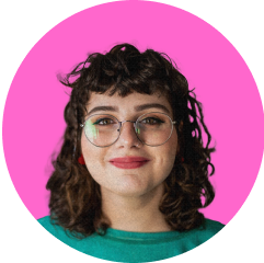 Smiling woman with curly hair, glasses, and a teal shirt against a bright pink background.